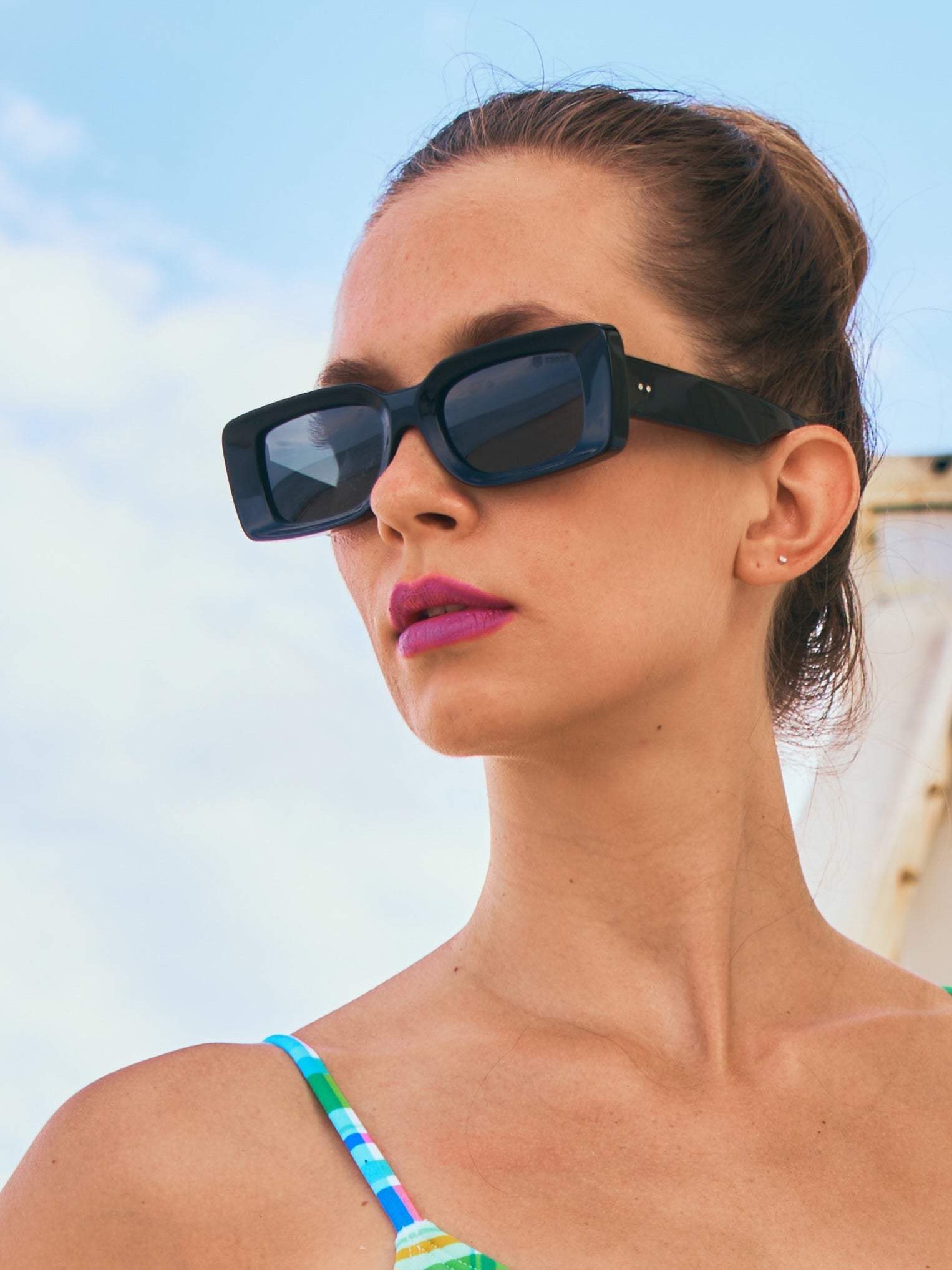 Woman modeling black rectangular sunglasses with bold frames and dark lenses, styled outdoors against a blue sky background.