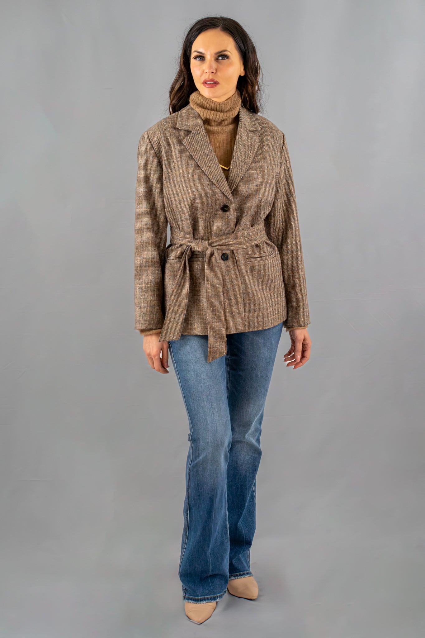 Woman wearing a brown belted blazer with a tailored fit, layered over a camel turtleneck sweater, paired with blue flared jeans and nude heels.