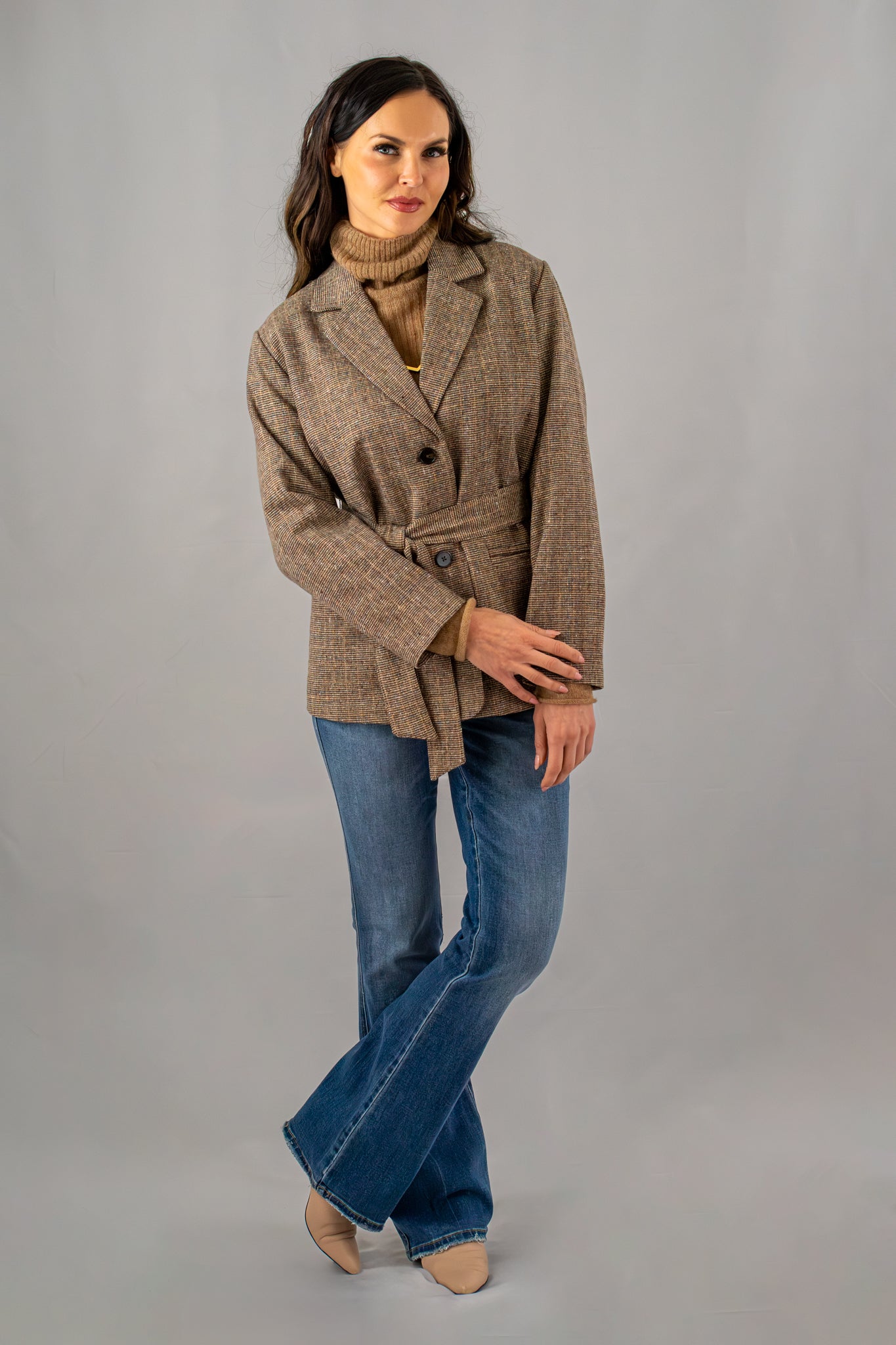 Woman styled in a brown belted blazer, camel turtleneck sweater, and blue flared jeans, showcasing a casual yet polished look.