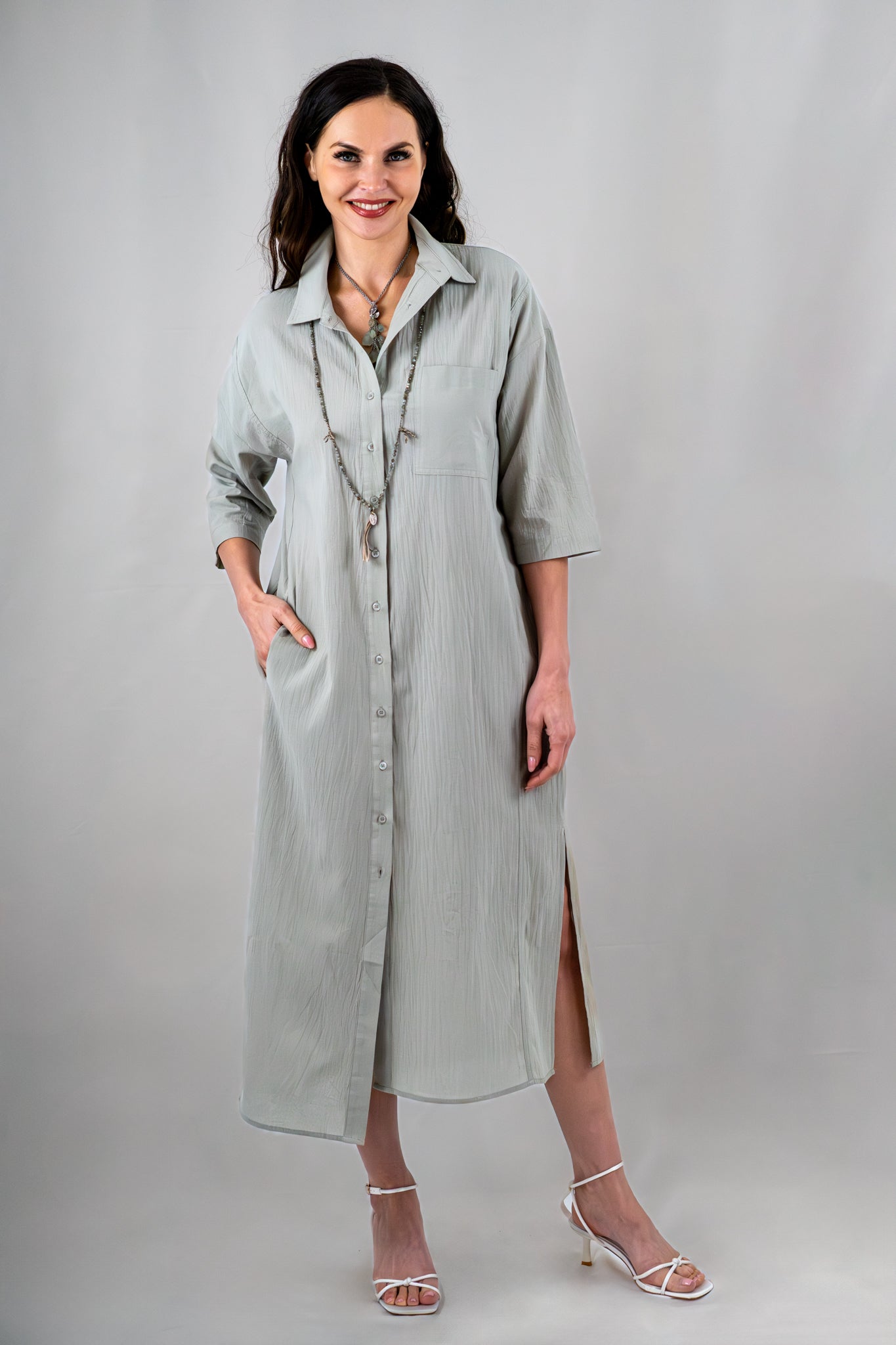 Woman wearing a sage green button-down midi dress with a collared neckline, side slits, and 3/4 sleeves, accessorized with layered necklaces and white strappy heels.