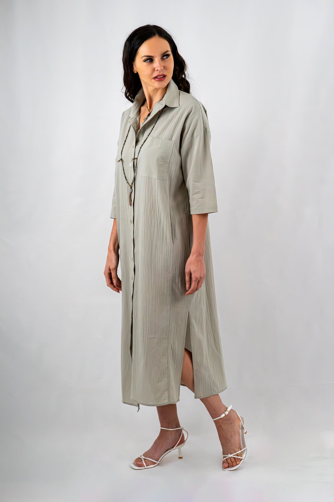 Side view of a sage green button-down midi dress with side slits and a collared neckline, styled with layered necklaces and white strappy sandals.