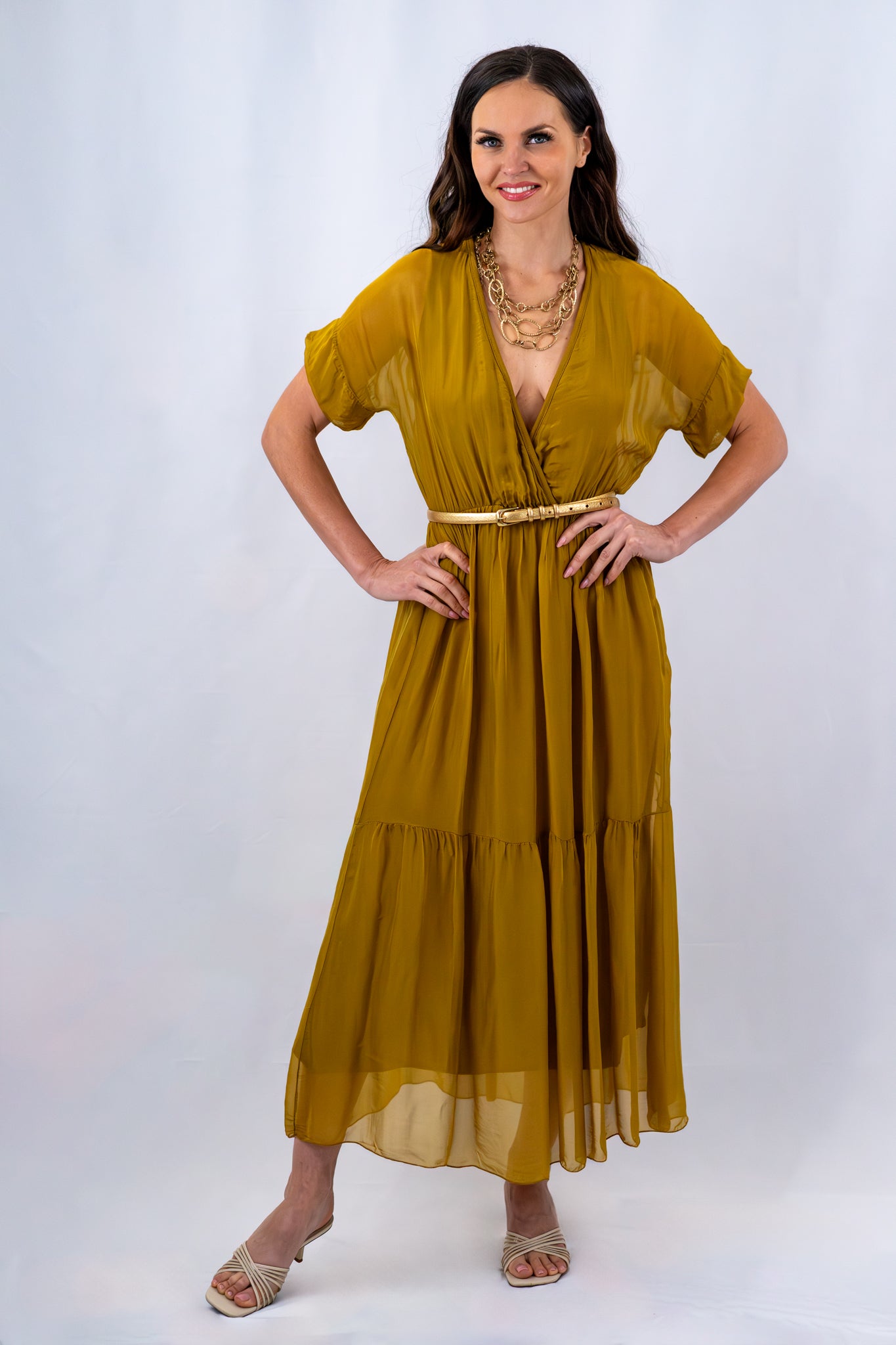 Woman wearing a flowy mustard yellow maxi dress with a gold belt and layered gold necklace, standing confidently in front of a plain white background, hands on hips.