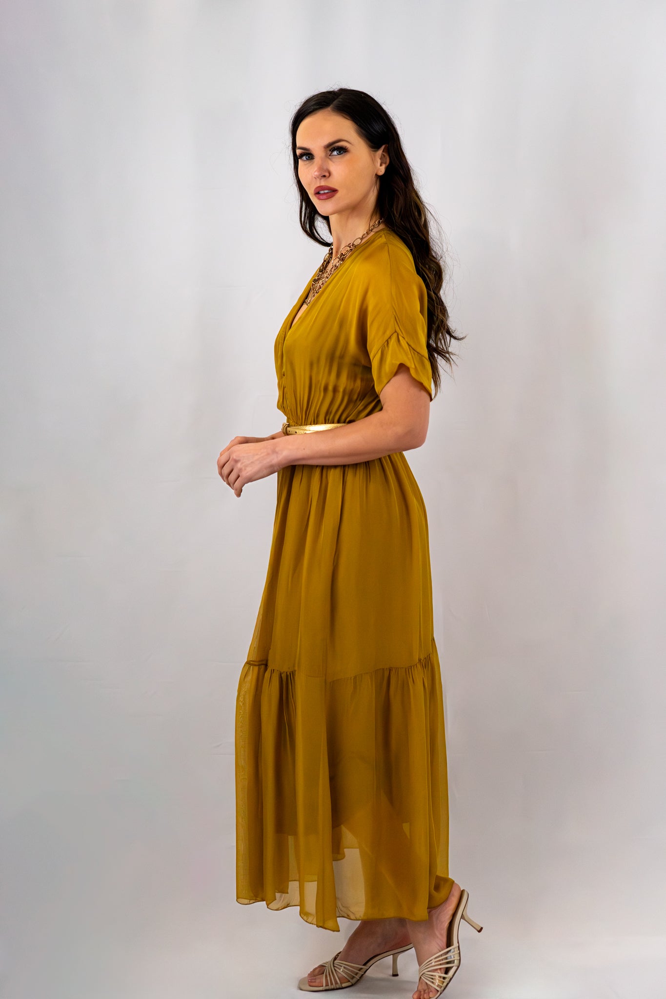 Woman in a mustard yellow maxi dress with a gold belt and layered necklace, captured in a side profile pose against a plain white background, showcasing the dress's silhouette.