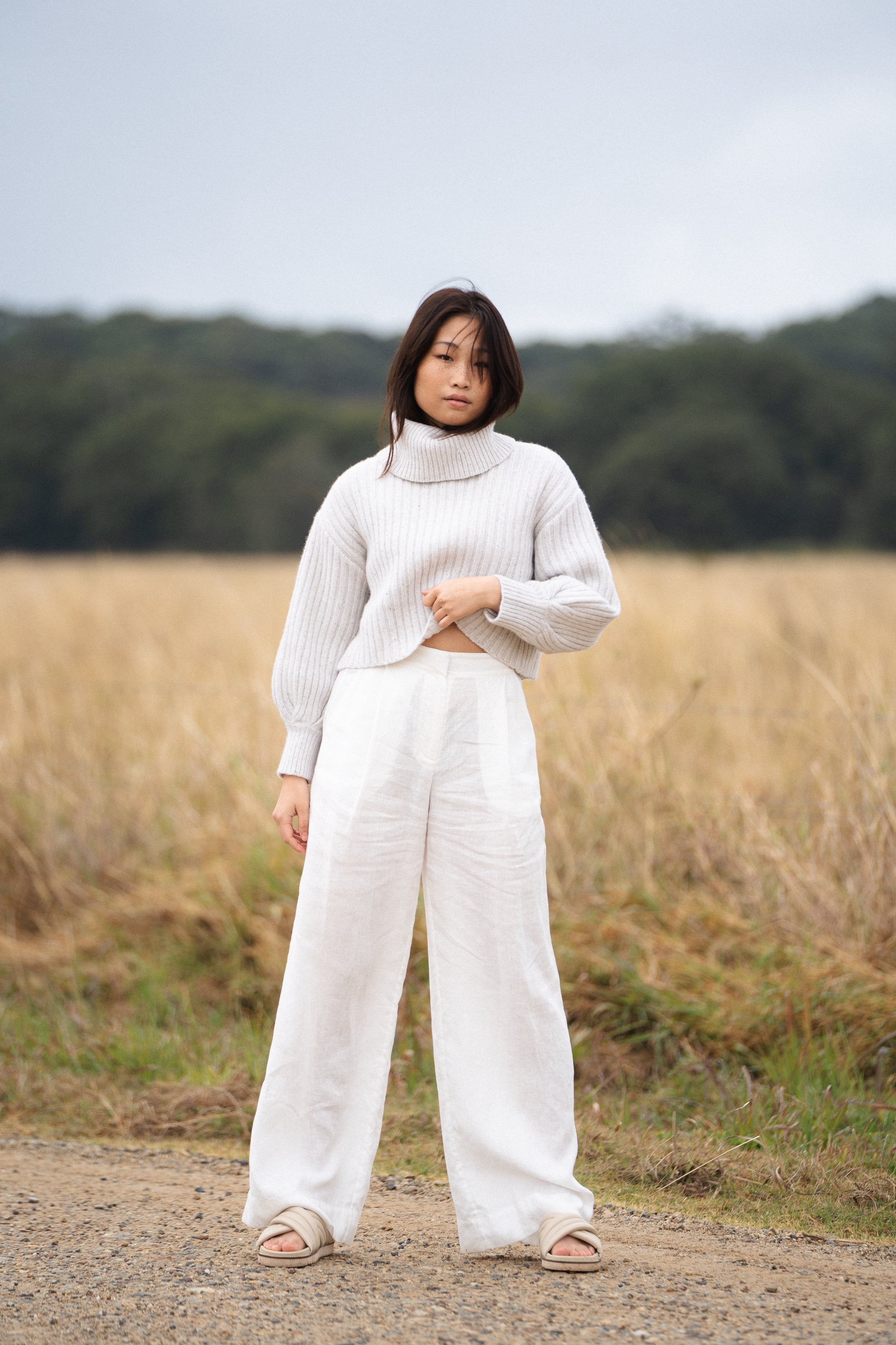 Woman wearing an oatmeal color ribbed turtleneck sweater, softly pulling the hem, standing outdoors with a golden field in the background.
