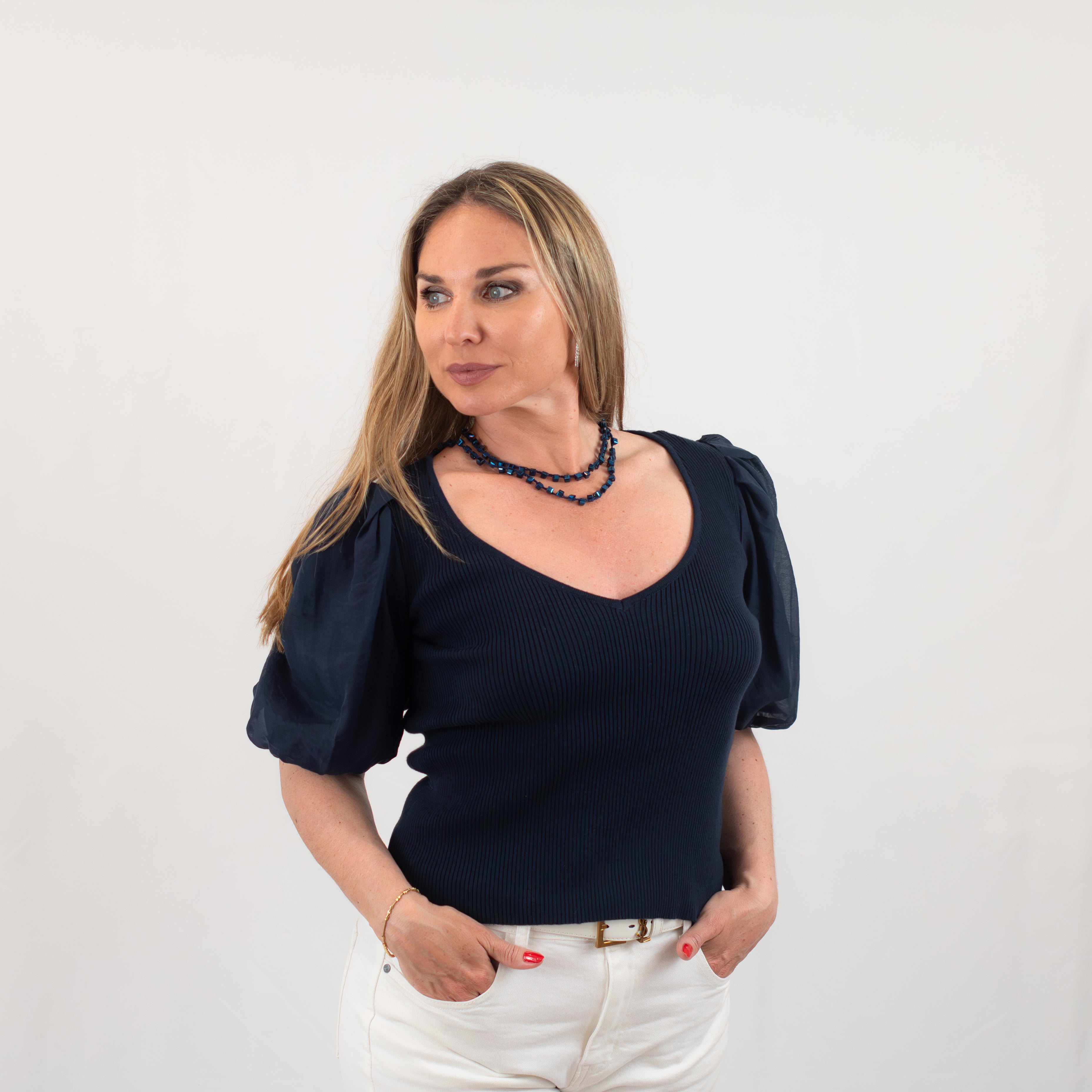 Side view of a woman in a navy puff-sleeve knit top with a V-neck, styled with white pants and accessorized with a layered necklace.