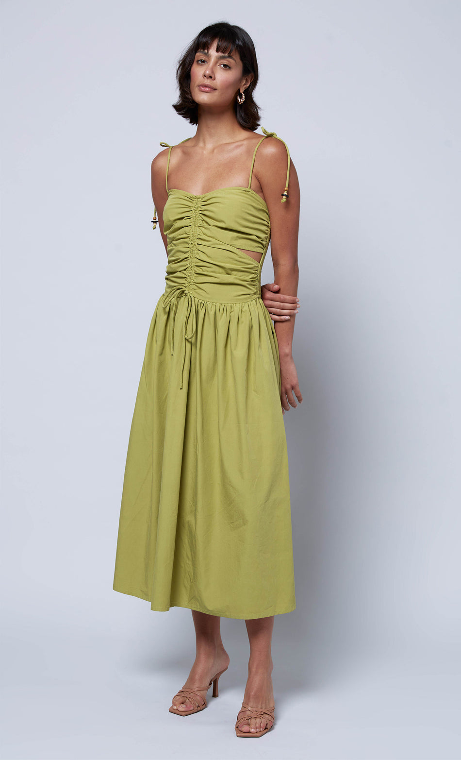 Woman modeling a green ruched midi dress with tie straps and cut-out details, styled with neutral heels for an elegant look.