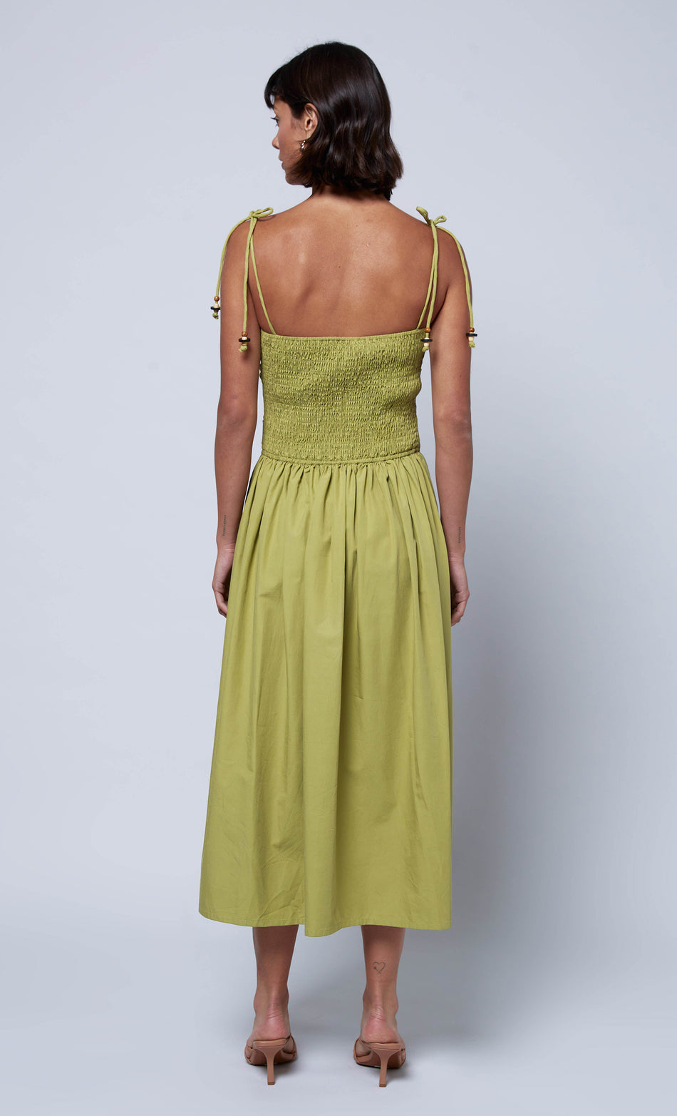 Back view of a green ruched midi dress featuring smocked detailing and adjustable tie straps, paired with neutral heels.