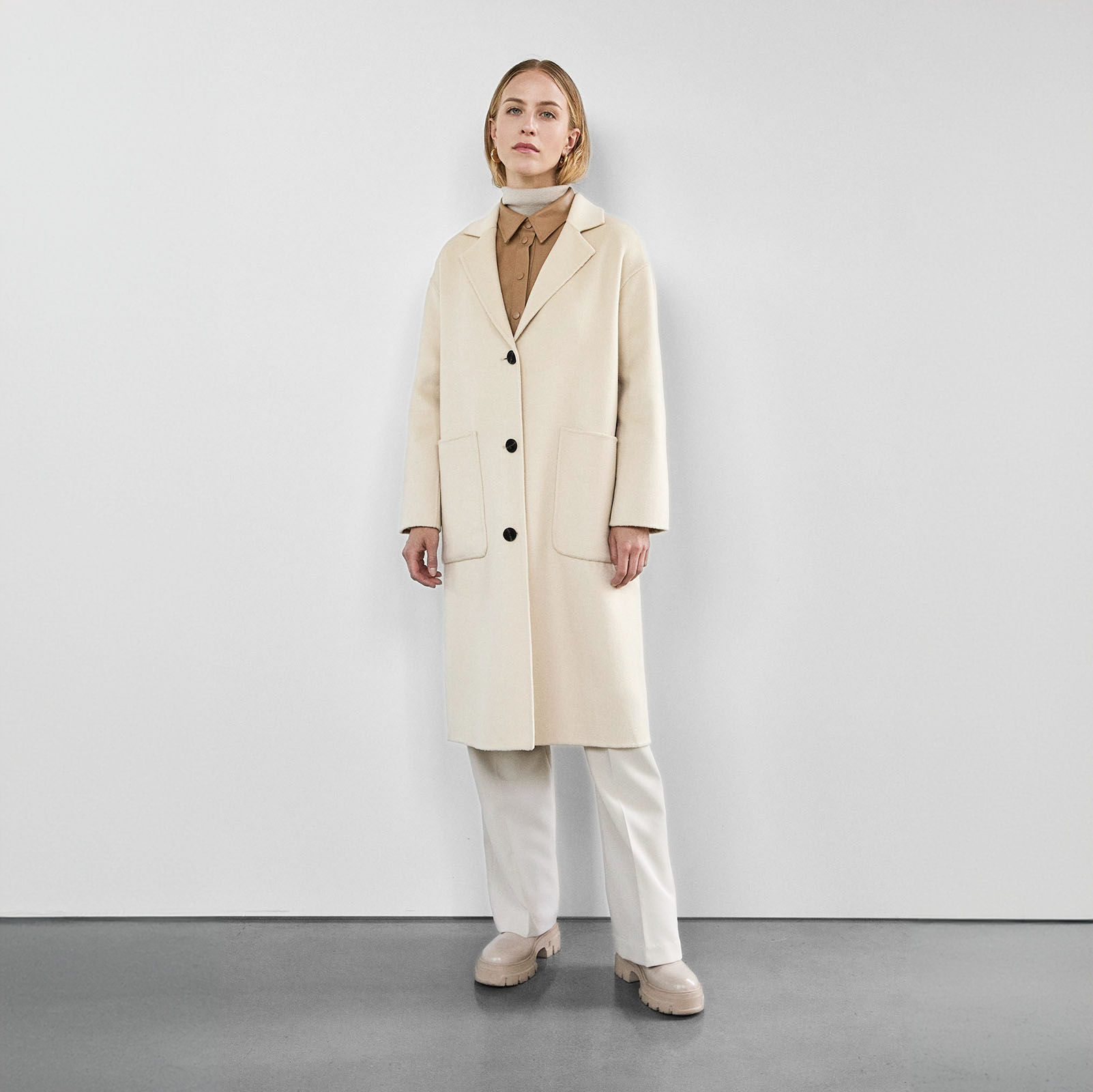 Woman wearing a cream wool coat with a classic lapel collar and black buttons, styled with a beige top, white trousers, and chunky beige boots.