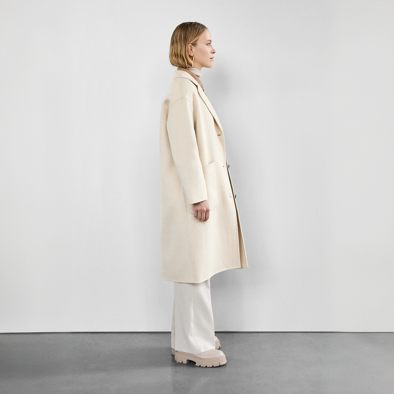  Side profile of a woman in a cream wool coat with large front pockets and black buttons, paired with white pants and beige boots.