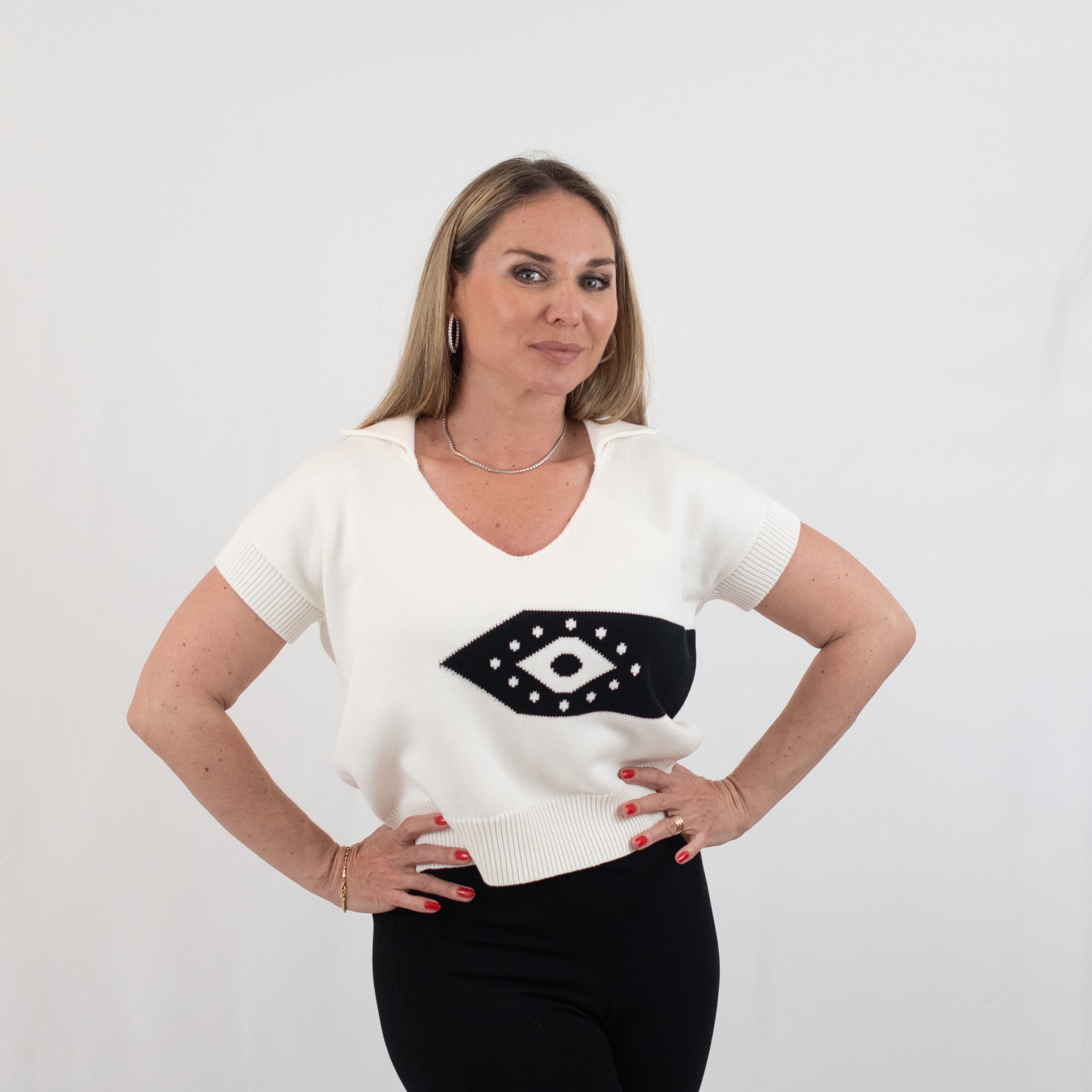 Woman wearing the Ada Short Sleeve Jumper in ivory, featuring a bold black eye motif with cream detailing on the front. This V-neck sweater, crafted from 100% organic cotton, has short sleeves and a ribbed hem, offering a chic and comfortable look suitable for casual and semi-formal occasions.