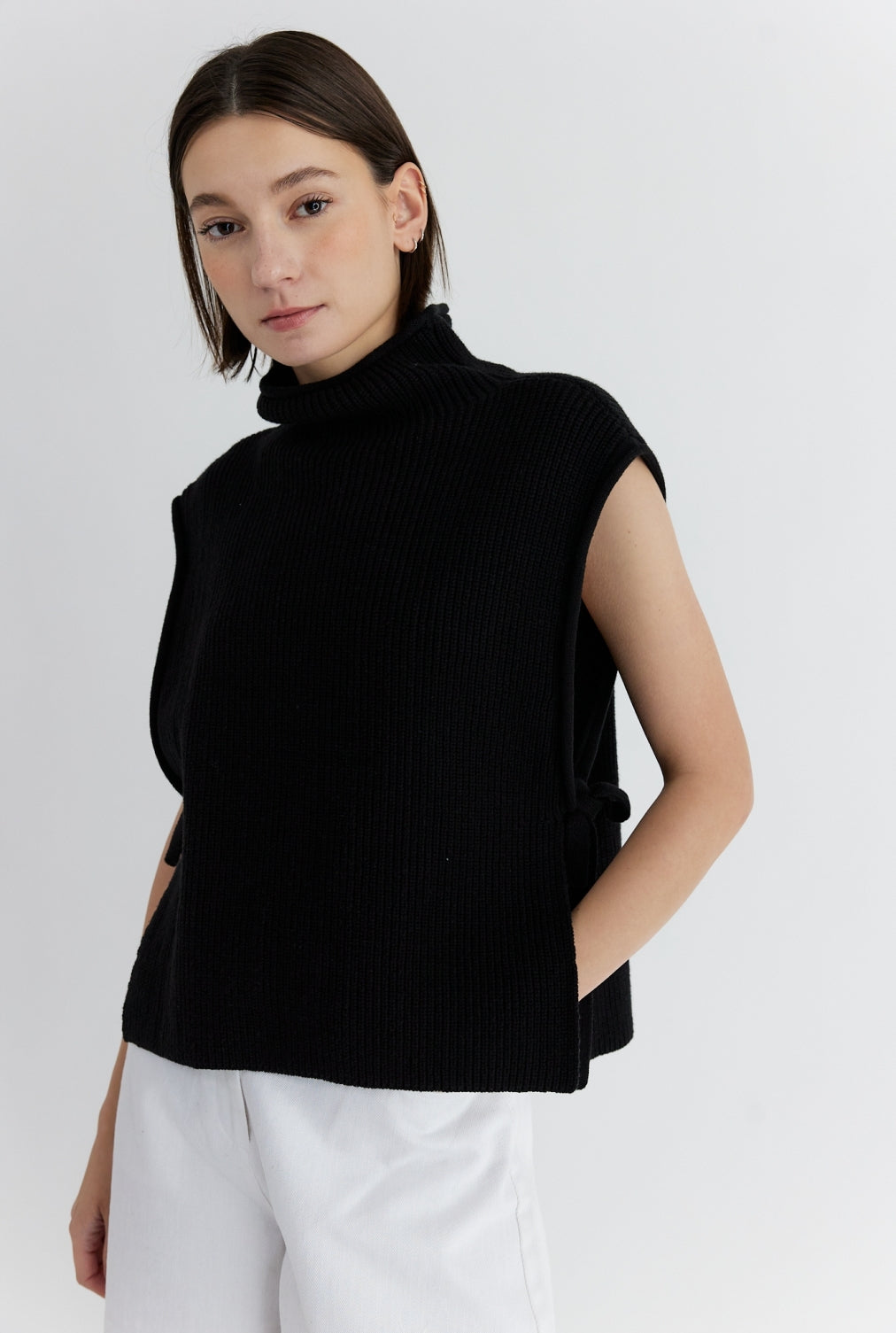 Woman wearing a black sleeveless turtleneck sweater with a ribbed texture and side ties, styled with white pants against a white background.