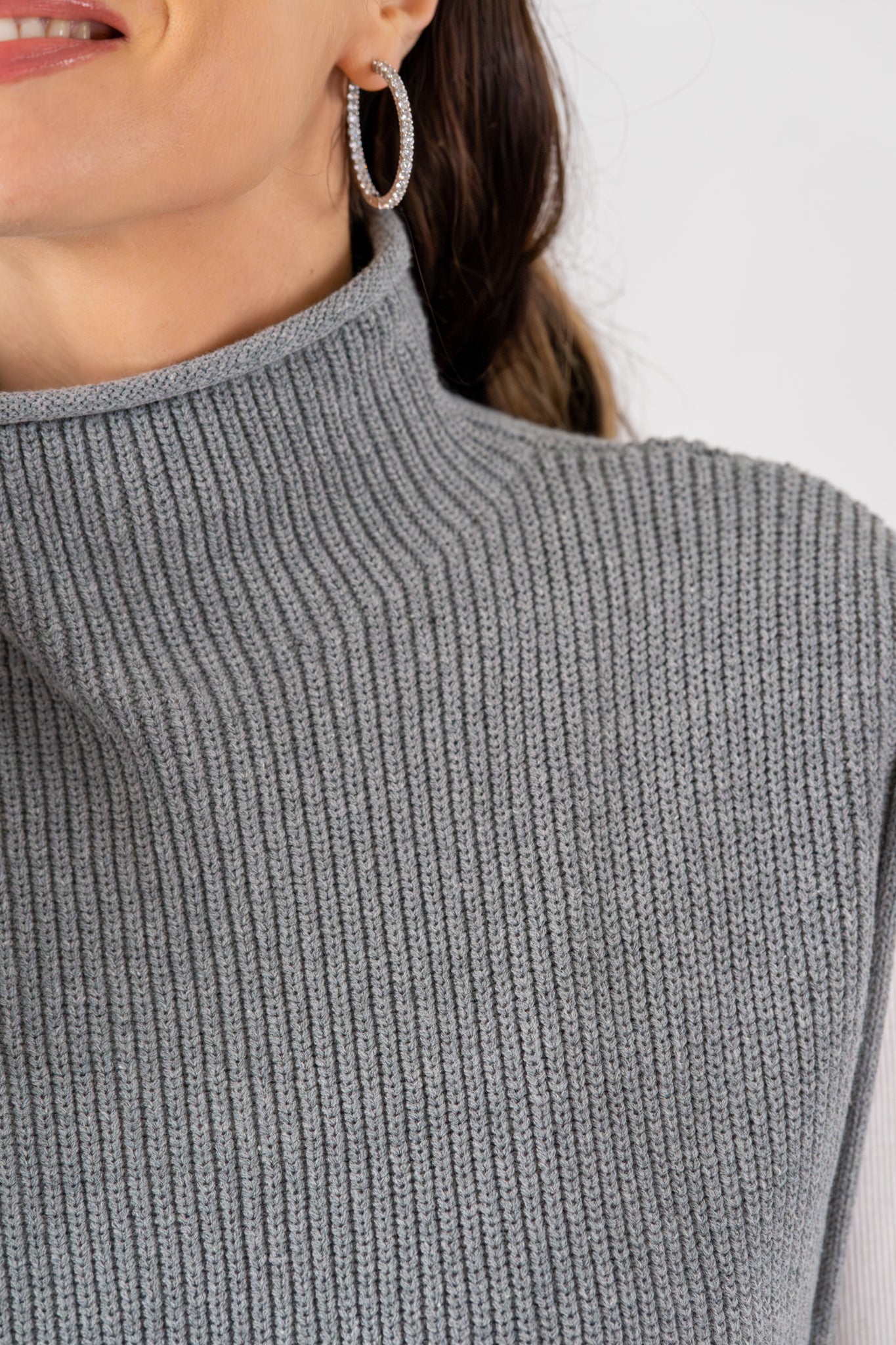 Close-up of a grey ribbed sleeveless turtleneck sweater worn by a woman, showing fabric texture and turtleneck detail with hoop earrings.