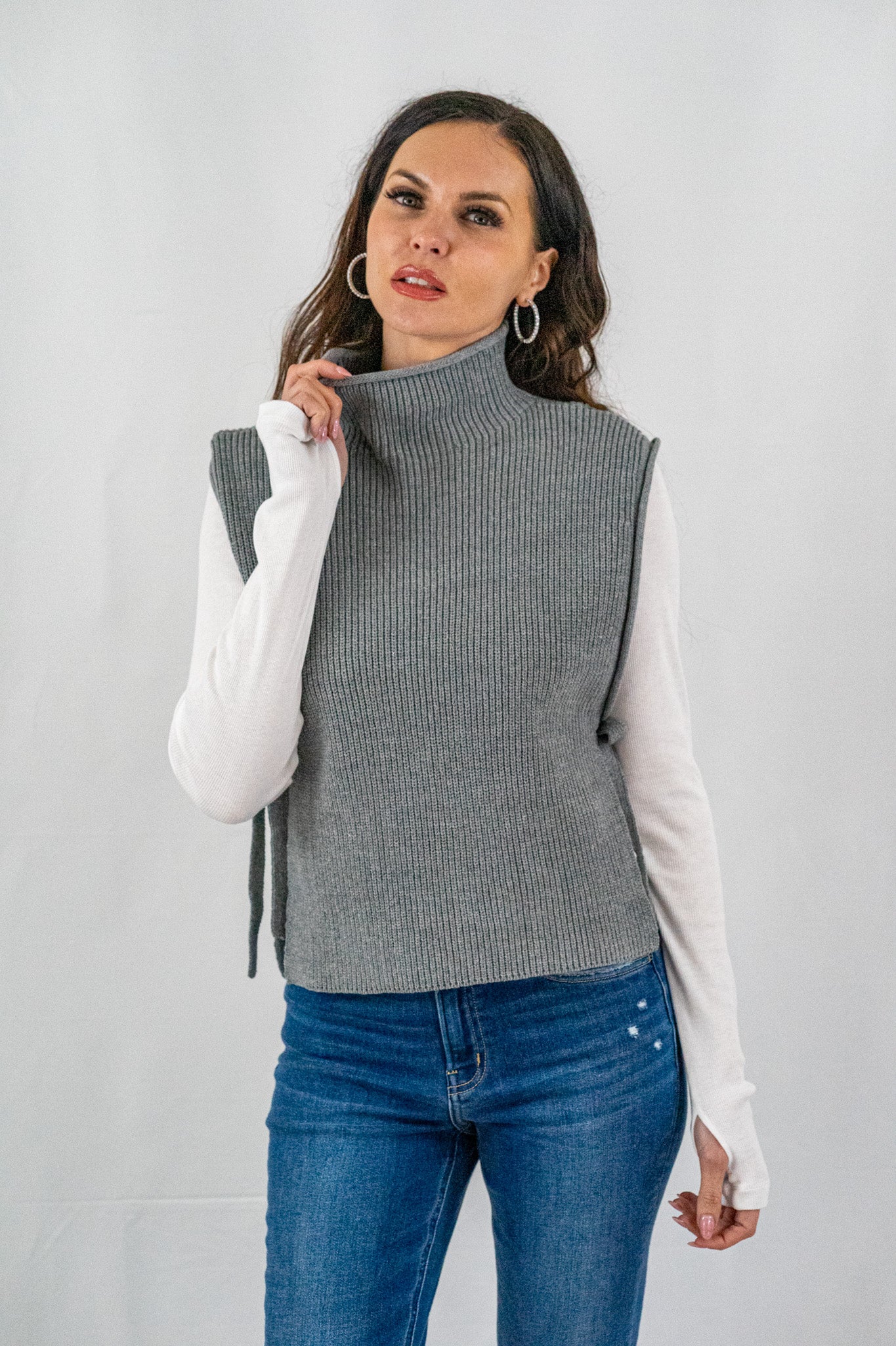 Woman wearing a grey sleeveless turtleneck sweater with a ribbed texture and side ties, layered over a white shirt with blue jeans, in a chic, casual style.