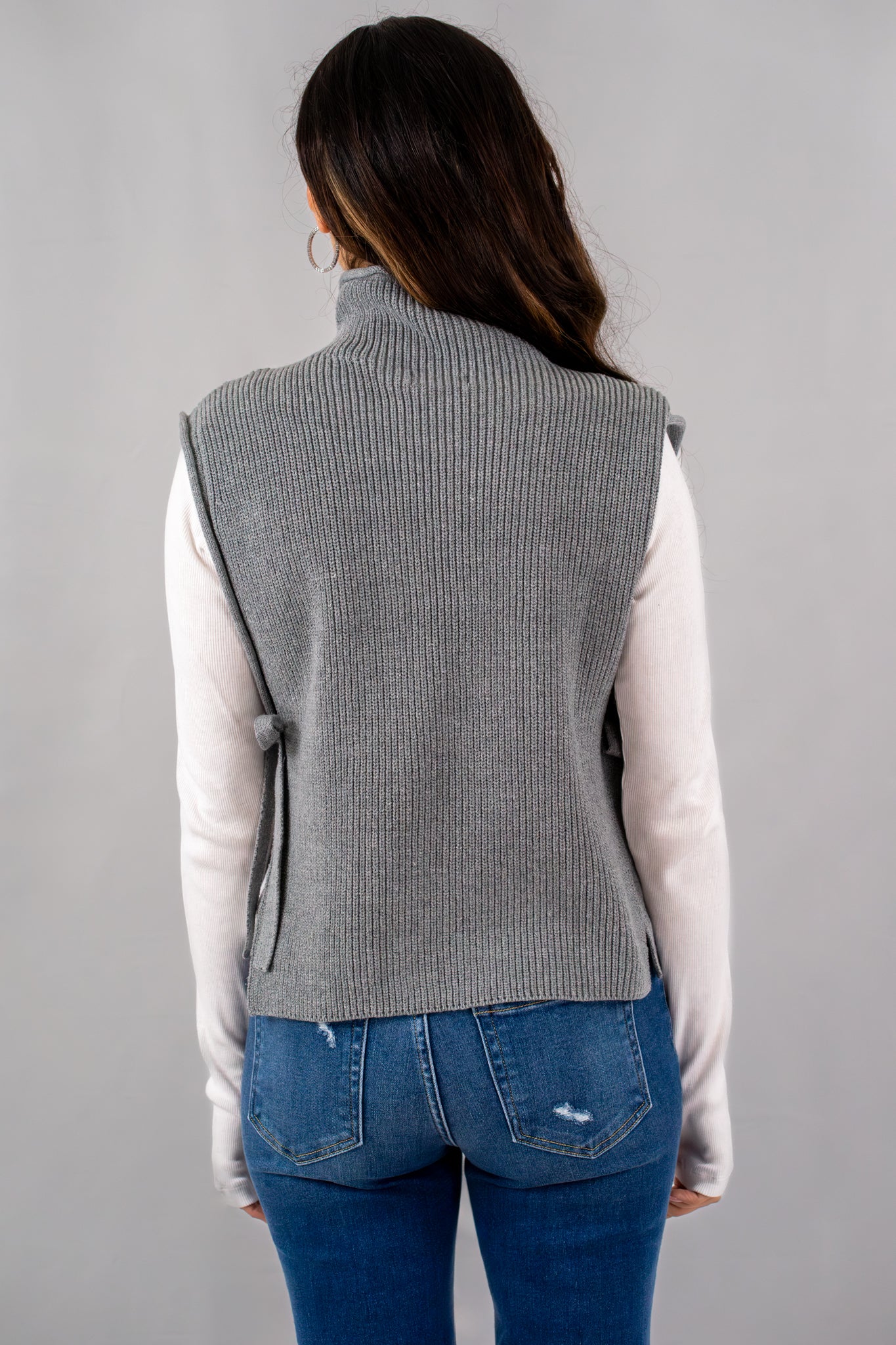 Back view of a grey sleeveless turtleneck sweater with side ties, worn over a white long-sleeve shirt and paired with blue jeans.