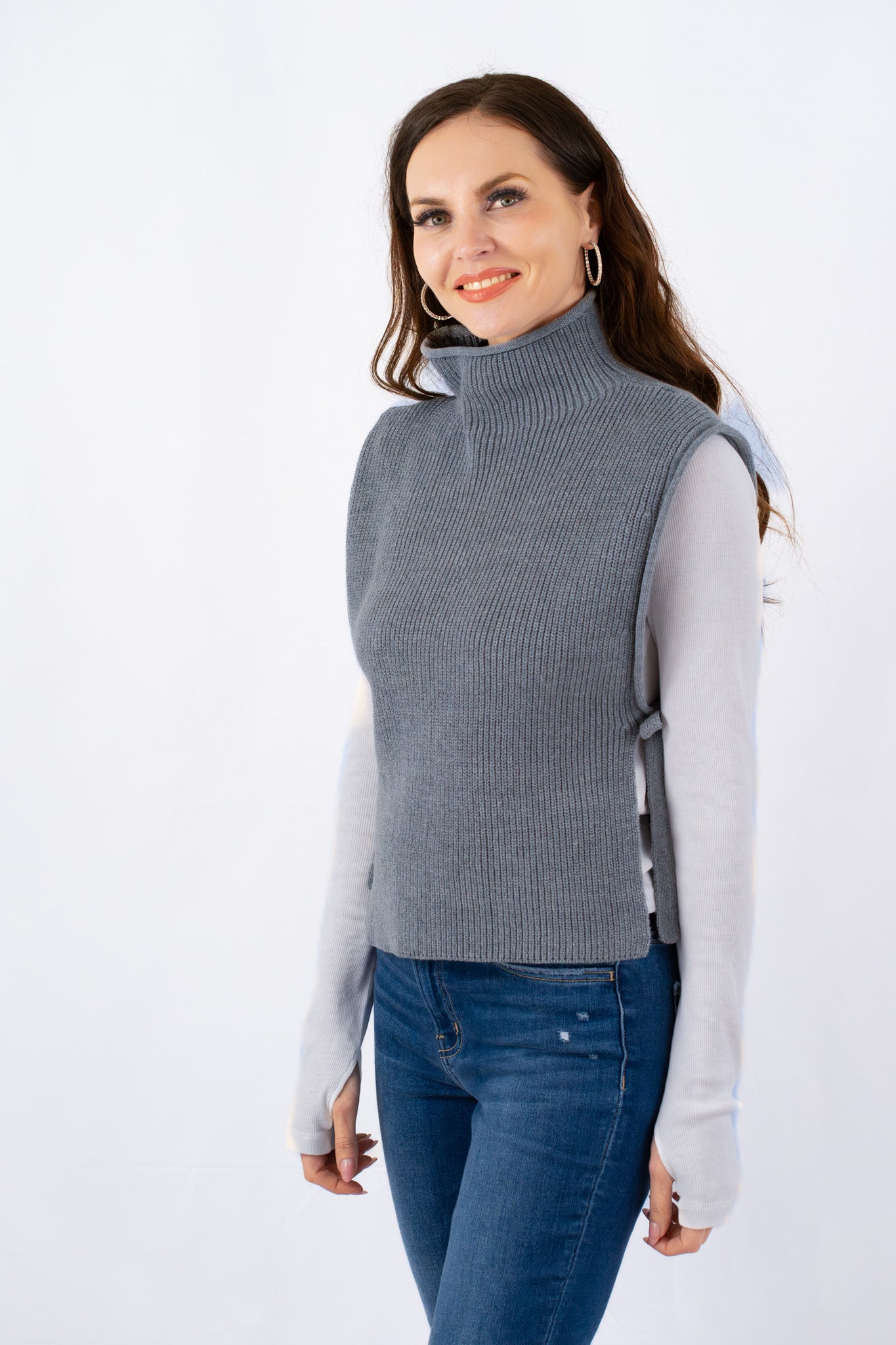 Woman wearing a grey sleeveless turtleneck sweater with side ties over a white long-sleeve shirt, styled with blue jeans.