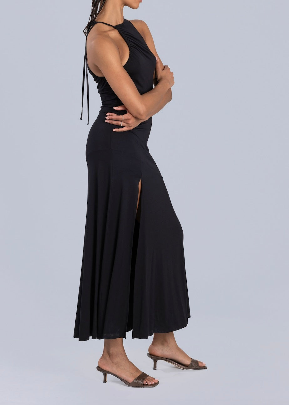 Side profile of a woman in a black halter-neck maxi dress featuring a thigh-high slit, styled with brown heeled sandals, set against a light gray background.