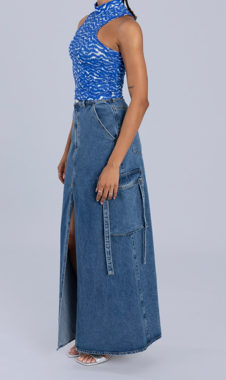 Side view of a blue maxi denim skirt featuring cargo pockets, a front slit, and casual detailing, styled with a blue patterned top.