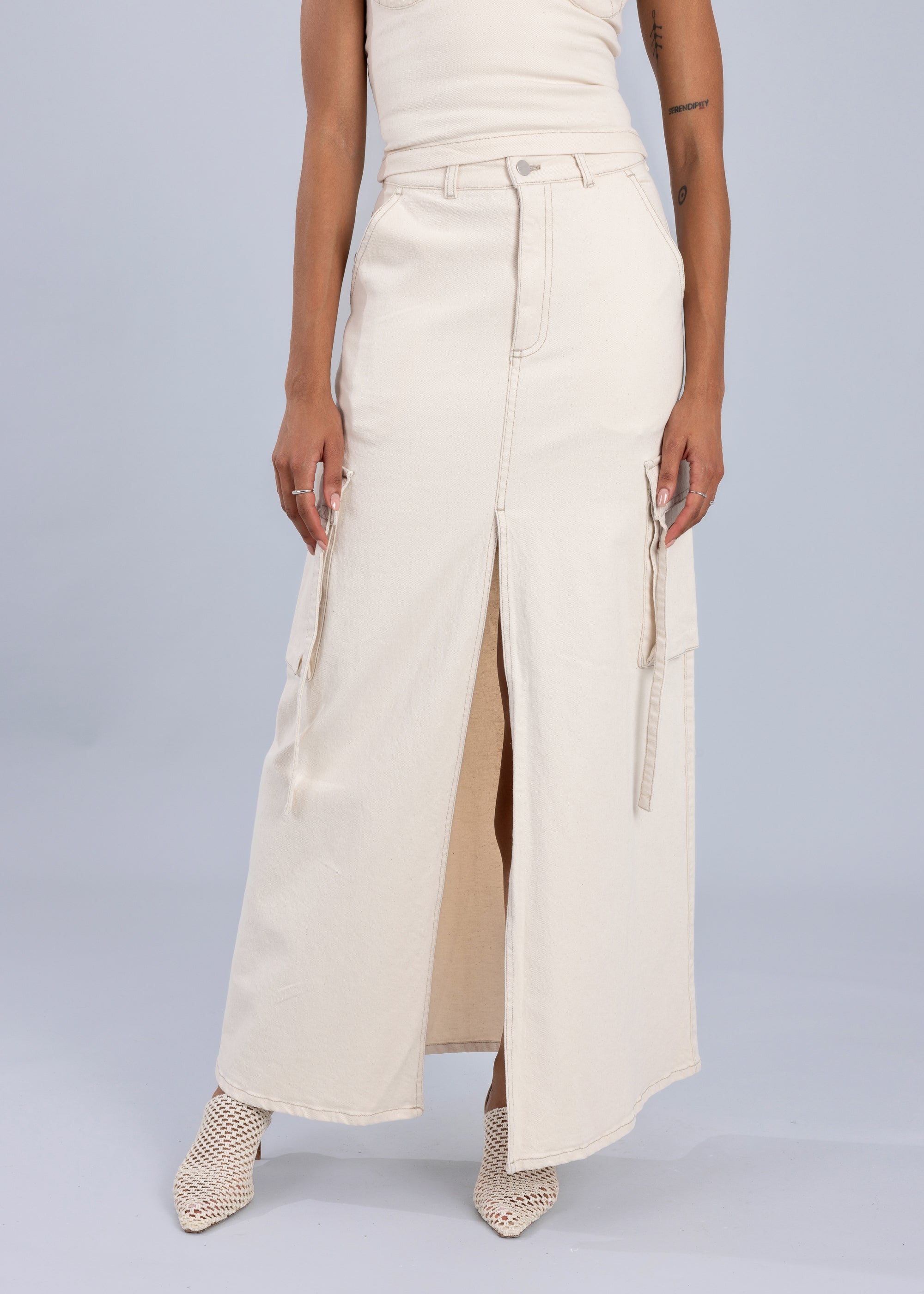 Woman wearing a high-waisted white maxi denim skirt with cargo pockets and a front slit, styled with a sleeveless cream top and woven heels.