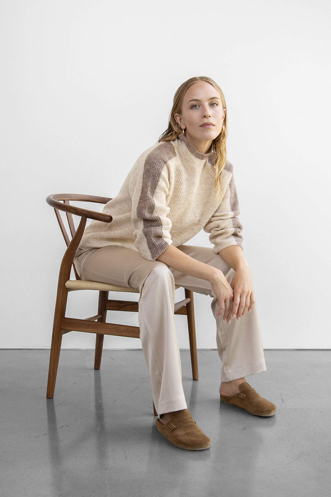 Woman seated in a wooden chair wearing a cream sweater with taupe accents, paired with beige pants and brown loafers for a relaxed, stylish look.