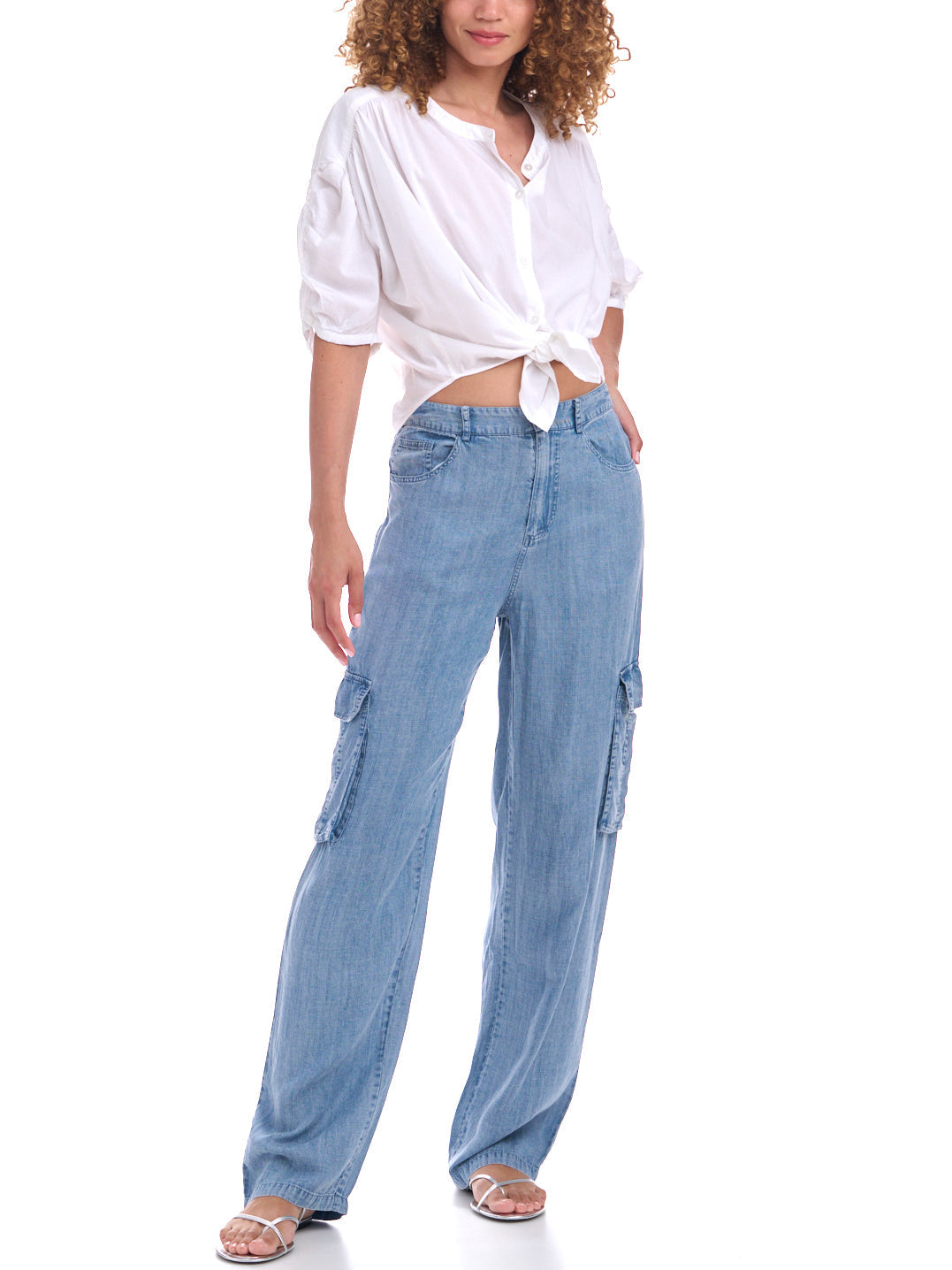 Woman wearing light-wash wide-leg cargo jeans with utility-style side pockets, paired with a white button-up shirt tied at the waist and strappy sandals.