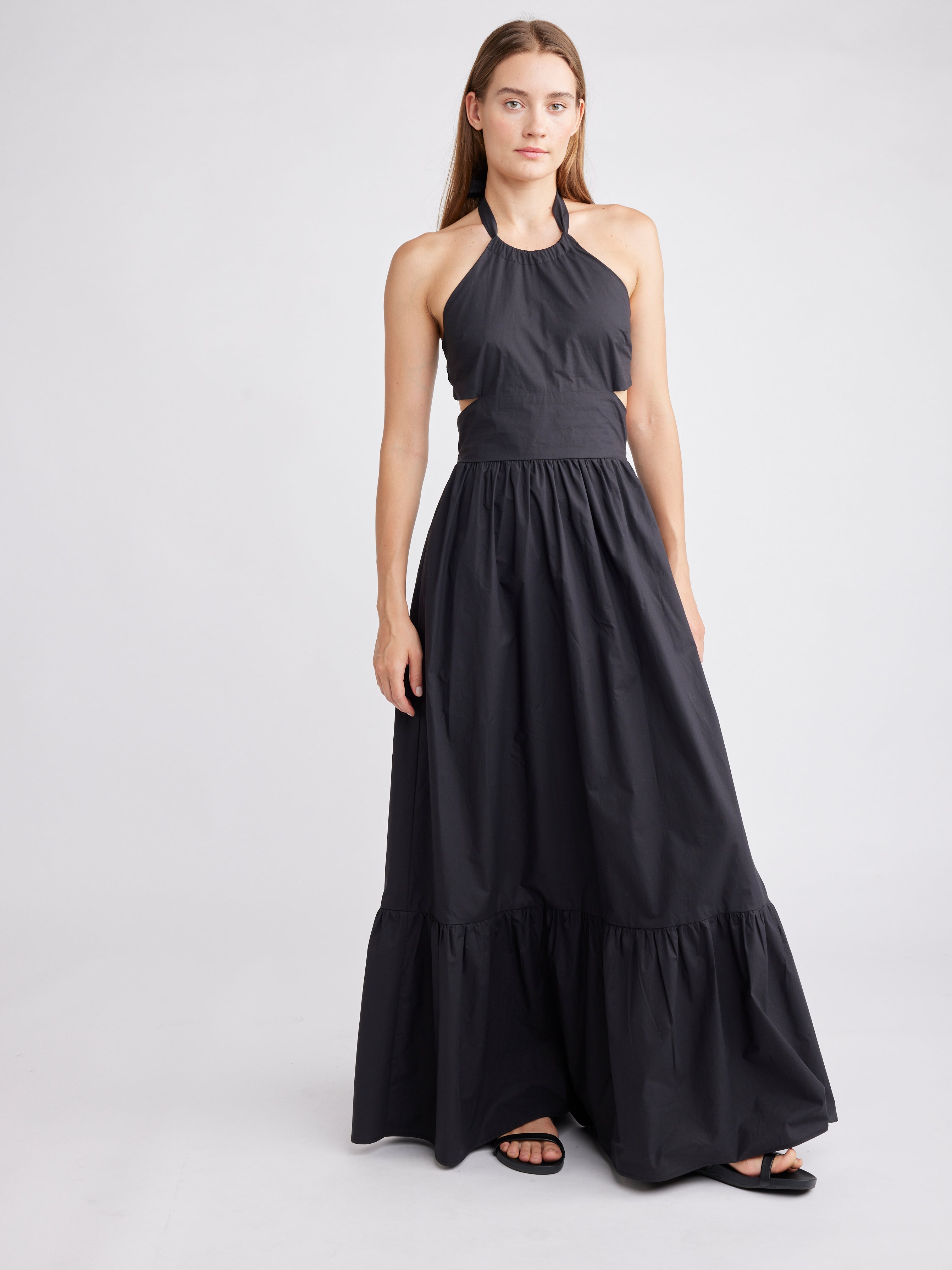 Model wearing an elegant black halter neck maxi dress with a tiered skirt, cinched waist, and sophisticated high neckline, paired with simple black sandals