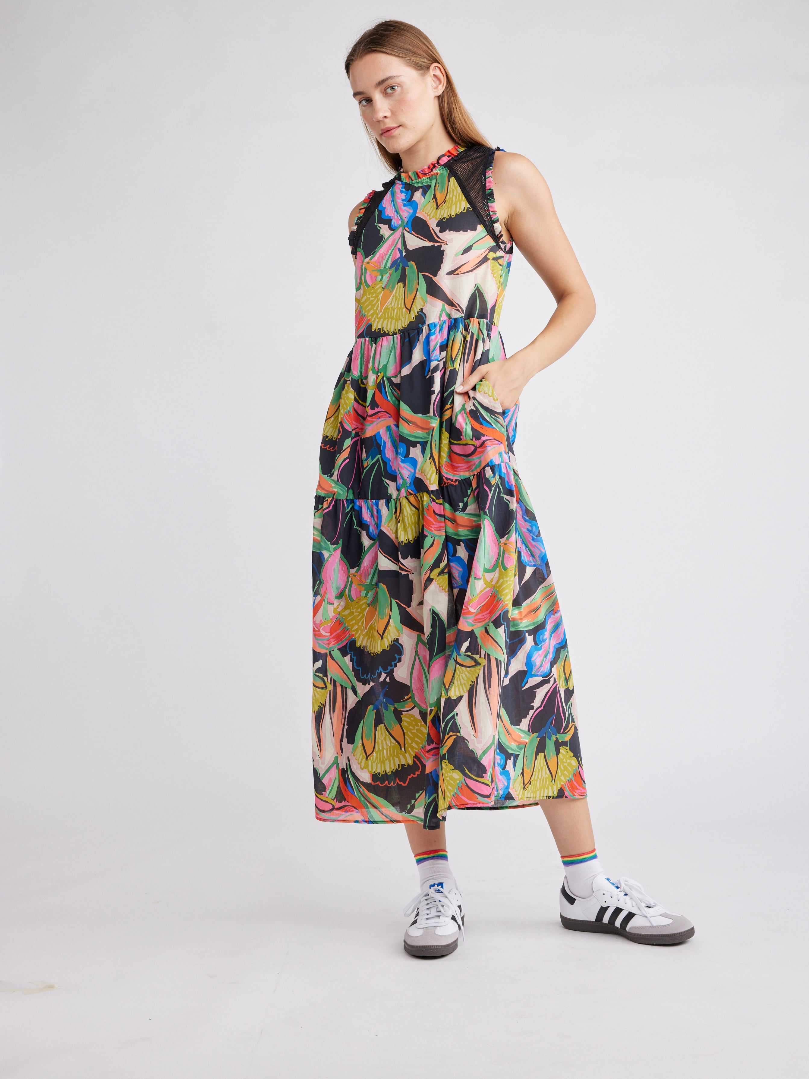 A model poses in a vibrant tropical print midi dress with a high neck, sleeveless design, cinched waist, and tiered skirt.