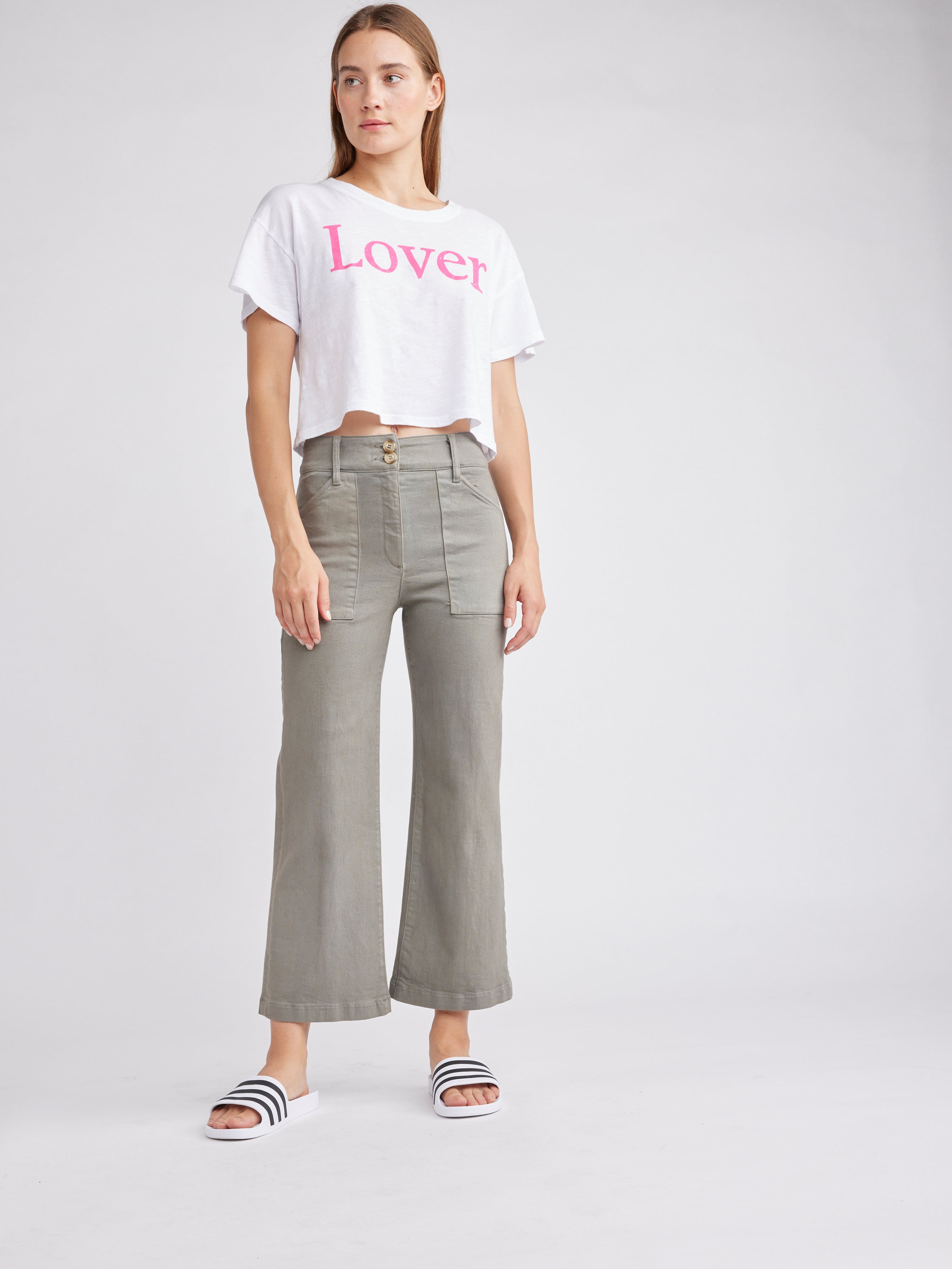 Woman wearing a cropped white t-shirt with "Lover" printed in pink, paired with olive green high-waisted flared pants and black-and-white striped slides, standing against a neutral background.