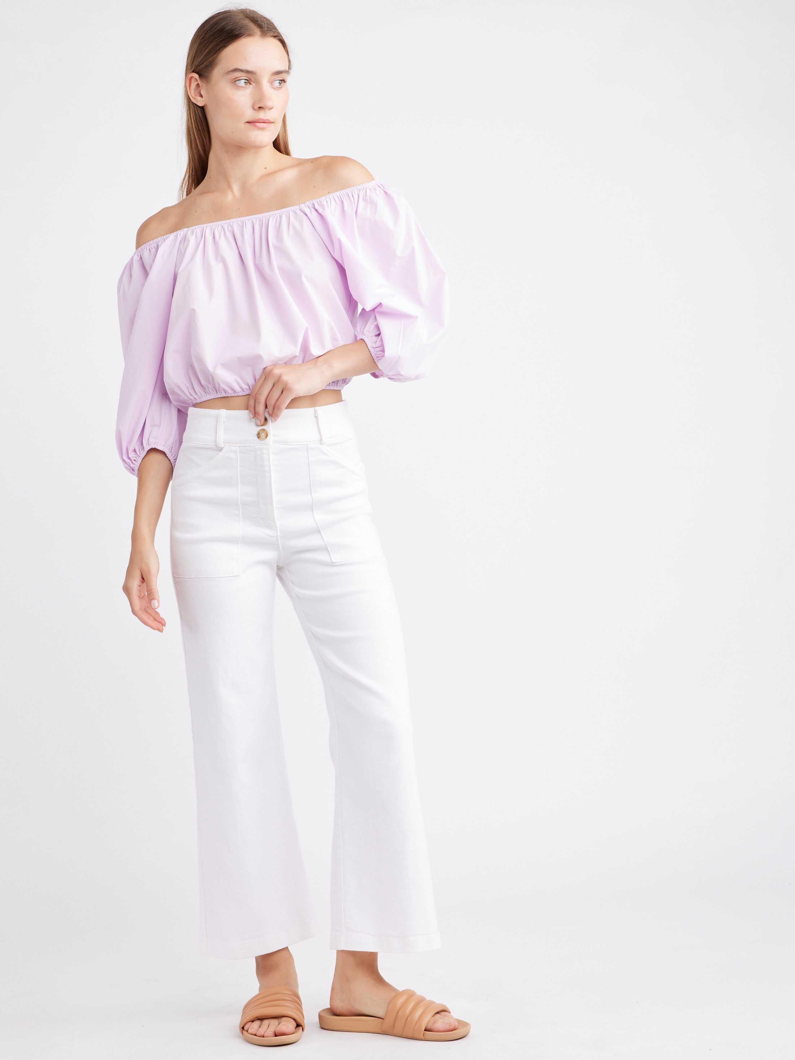 Woman dressed in a lilac off-shoulder puffed-sleeve top paired with white high-waisted flared pants and tan slide sandals, standing against a plain white background.