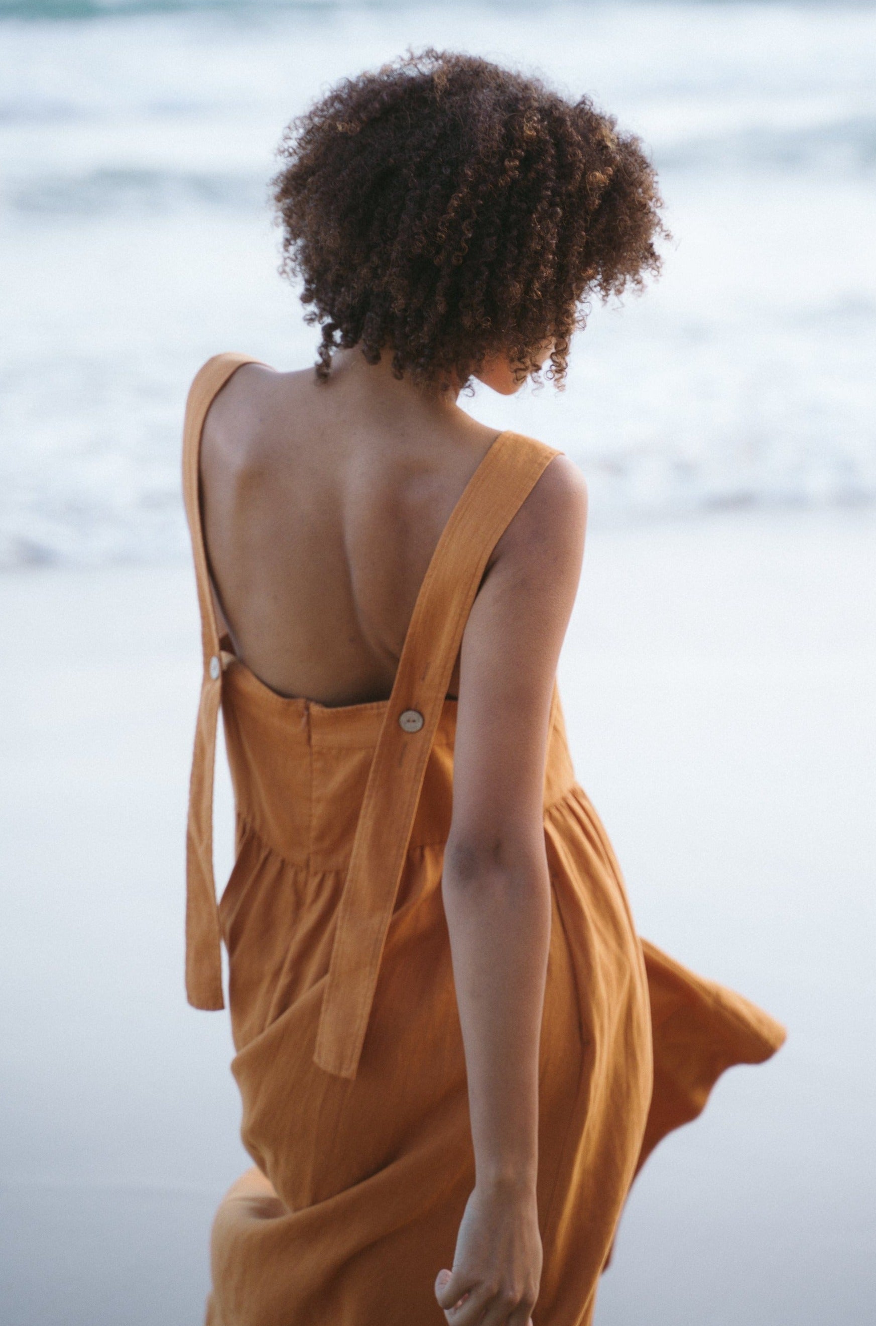 Rear view of the Frida Linen Dress highlighting the cross-back straps with button details at the waistline and the elegant high-low hemline.