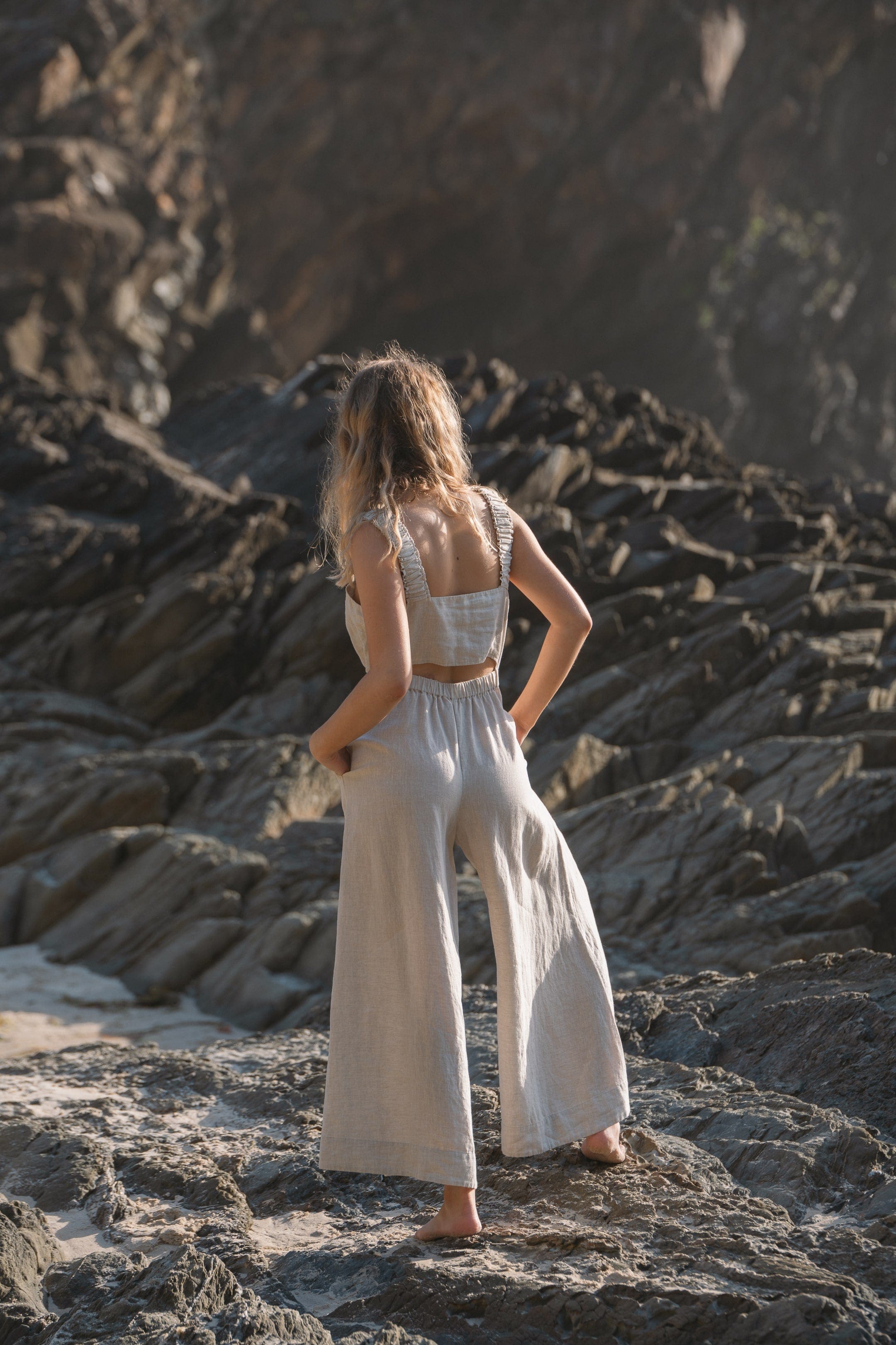 Back view of a sustainable oatmeal linen jumpsuit featuring an elegant back cutout, elasticized waist, and flowing wide-leg design on a rocky outdoor backdrop.